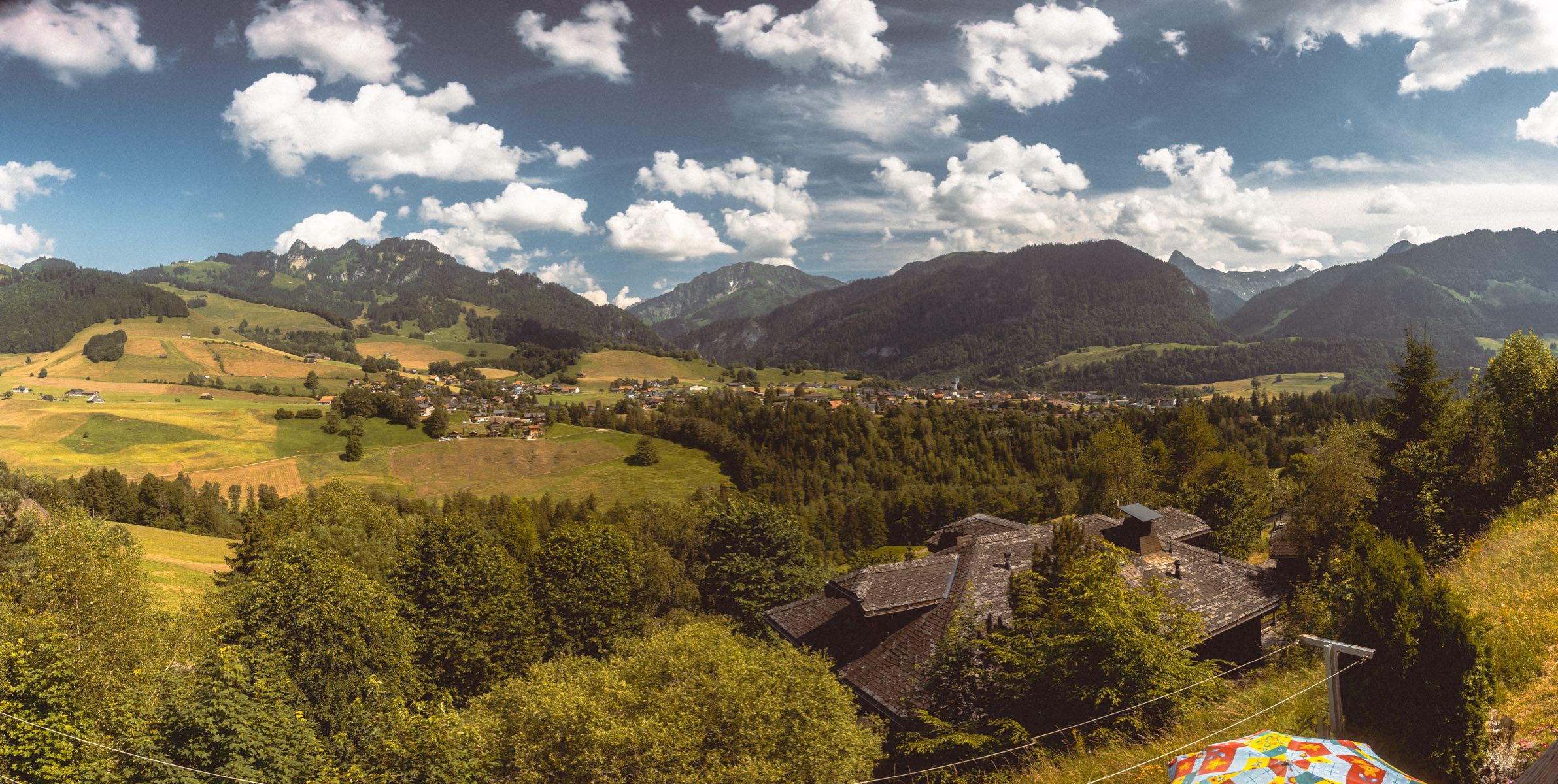 A wide summer view from the chalet over the green valley: meadows and forest falling to a village with its church steeple, the Gruyere pre-Alps ranged across the horizon under a bright sky, the chalet's own slate roof among the trees at the lower right.