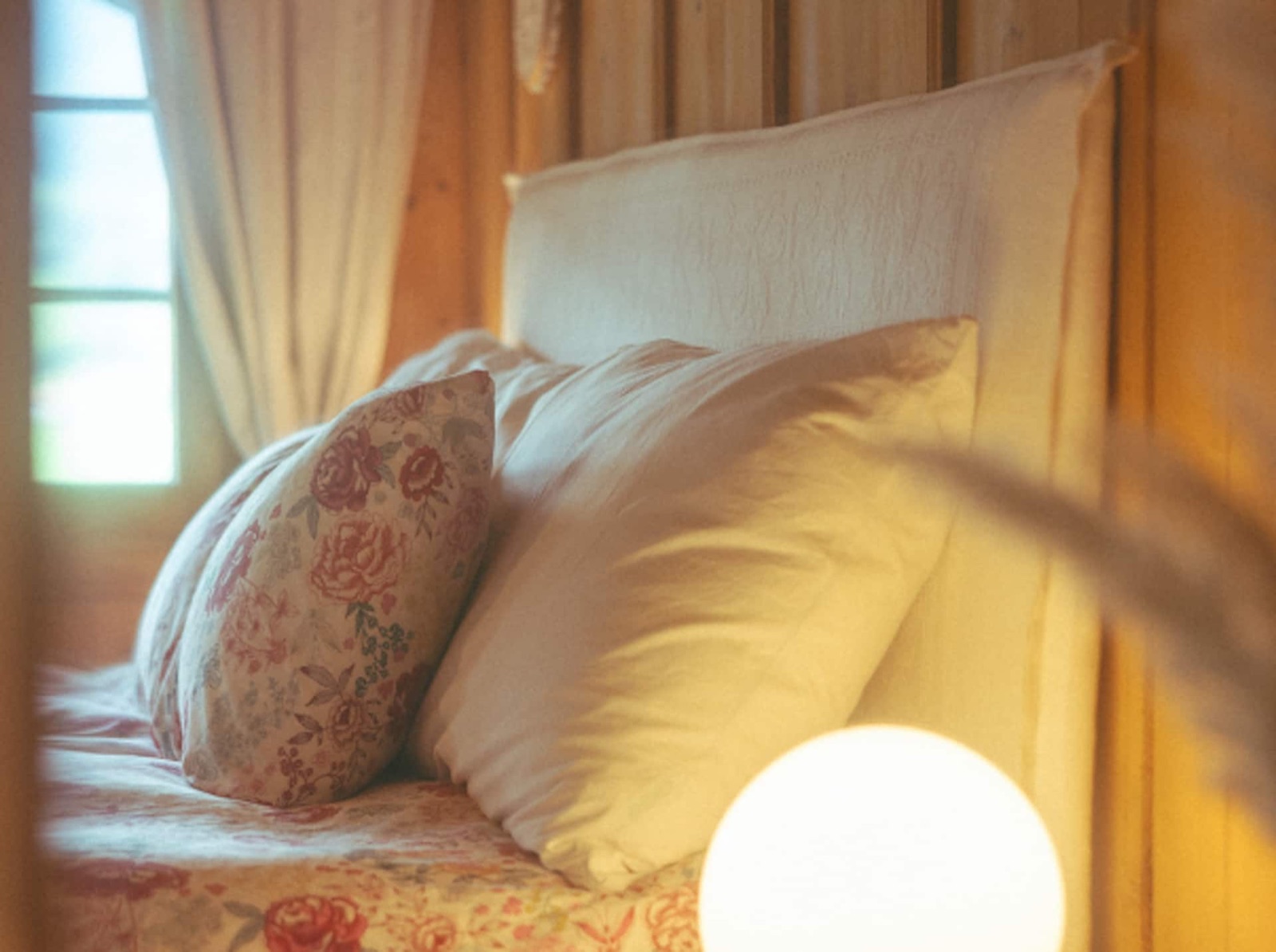A double bed made up in white cotton linens with a floral cushion, warm morning light through a linen curtain.